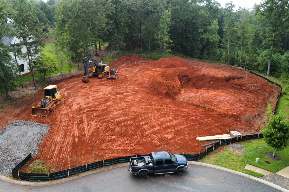 Aerial view of a cleared and graded homesite lot in a Georgia neighborhood with an excavator, dozer, and silt fence